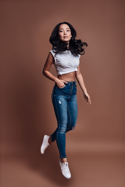 Playful beauty. Full length of beautiful young woman smiling and looking at camera while hovering against brown background