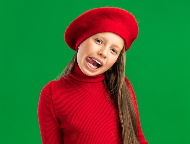 Playful little blonde girl wearing red beret showing tongue  isolated on green wall