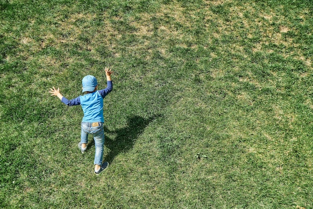 Playful little boy goes up steep hill slope covered with lush green grass in park backside view