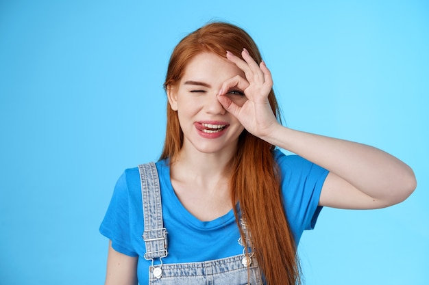 Playful lovely redhead female long red hairstyle, look through fingers carefree joyful, smiling show tongue sassy, enjoy summer vacation, stand blue background, tourist relax resort