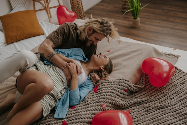 Playful loving couple embracing and having fun while sitting on bed surrounded with red balloons