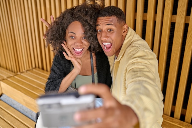 Playful mood. Dark-skinned young cheerful man and pretty woman with open mouth smiling taking selfie at camera sitting outdoors in afternoon