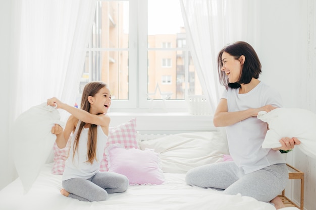 Playful mother and daugher have fun together, have pillow fight on bed, spend free time at home