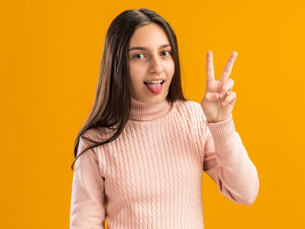 Playful pretty teenage girl  making peace sign showing tongue isolated on orange wall