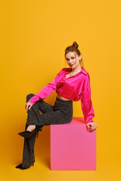 Playful woman in bright oldfashioned clothes sits in profile on purple cube over bright yellow studio background