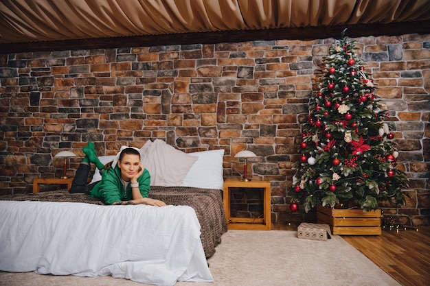 Photo playful woman in warm clothes laying on bed with crossed legs near the christmas tree