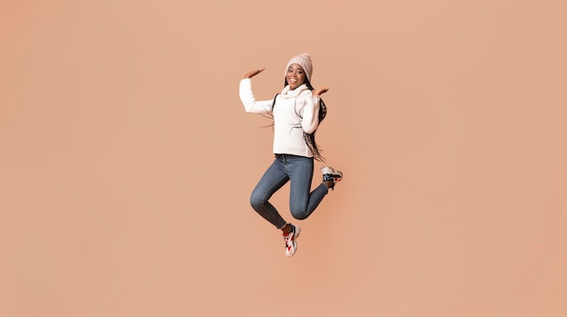 Photo playful young african woman in winter hat having fun over beige studio background jumping in the air