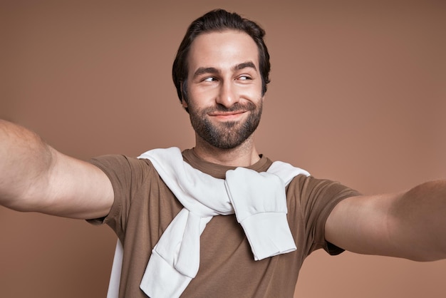 Playful young man making selfie and grimacing while standing against brown background