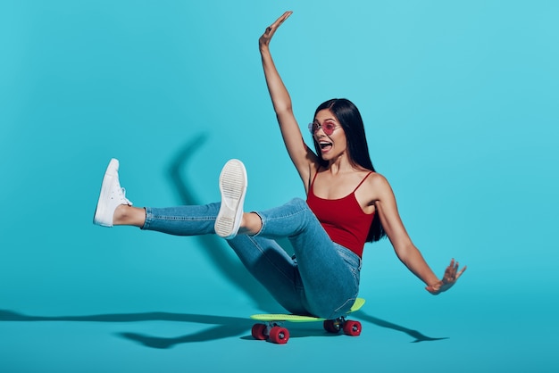 Playful young woman smiling while skateboarding against blue wall