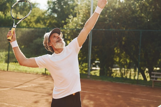 Playing game Senior modern stylish man with racket outdoors on tennis court at daytime