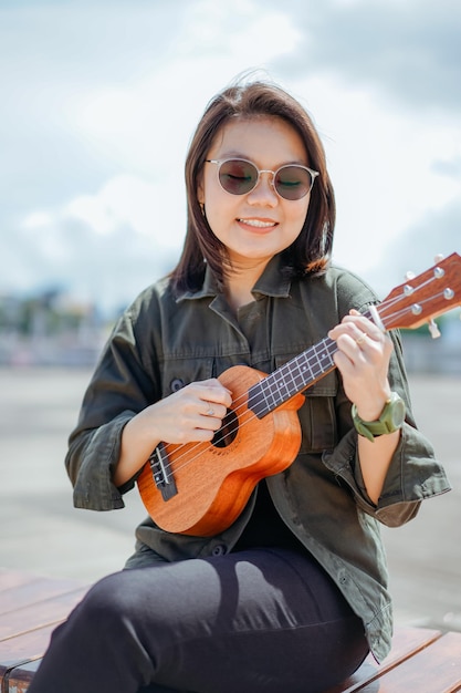 Playing Ukulele of Young Beautiful Asian Woman Wearing Jacket And Black Jeans Posing Outdoors