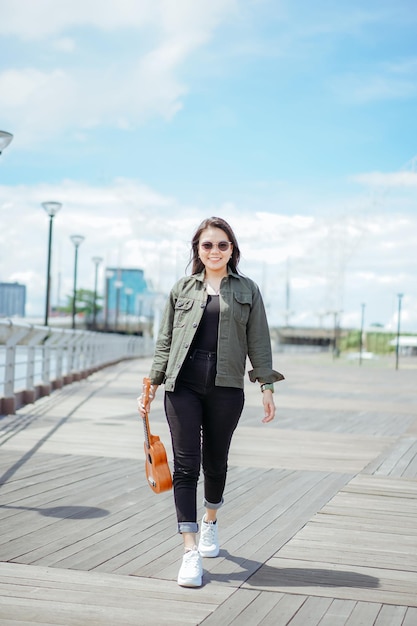 Playing Ukulele of Young Beautiful Asian Woman Wearing Jacket And Black Jeans Posing Outdoors