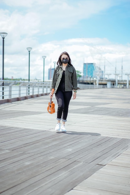 Playing Ukulele of Young Beautiful Asian Woman Wearing Jacket And Black Jeans Posing Outdoors