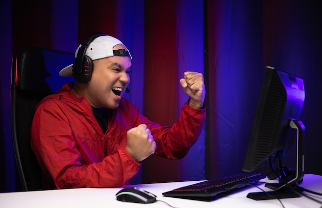 Playing video game. Young asian handsome man sitting on chair in living room. Happiness Streamer Indian man  playing game online wearing headphone in the darkroom.