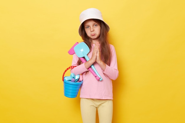 Pleading little cute girl in panama holding sandbox toys rake and shovel isolated over yellow background standing with paying gesture asking play at beack more