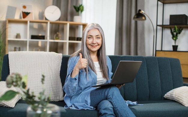 Pleasant attractive greyhaired woman with long hair sitting on couch with laptop on knees