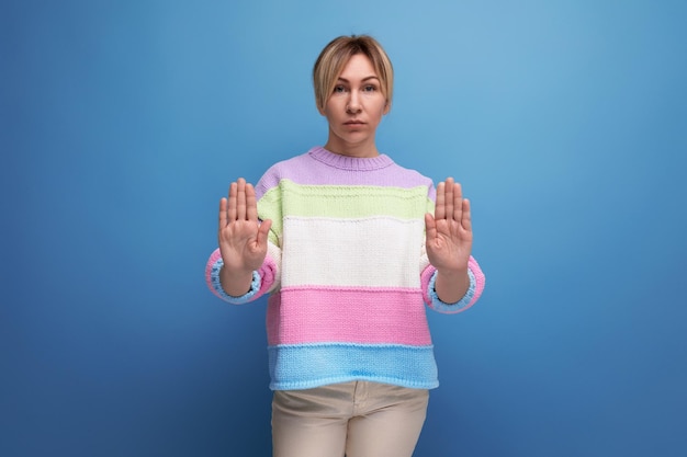 Pleasant blond woman in casual outfit showing stop sign on blue background with copy space