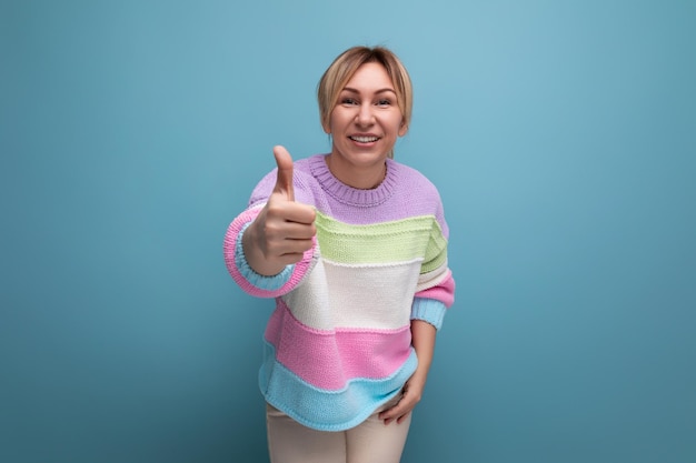 Pleasant blond young woman in a striped sweater shows a super gesture on a blue background