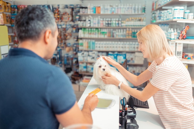 Pleasant cashier touching white dog while man paying by card