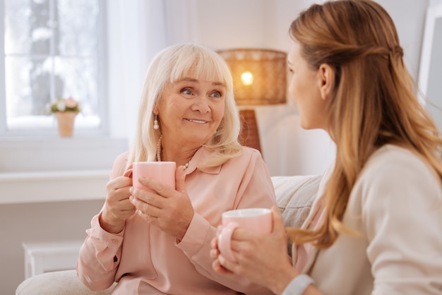 Pleasant conversation. Nice joyful elderly woman holding a cup and smiling while talking to her daughter