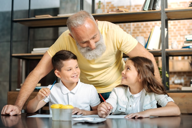 Pleasant conversation. Sweet pre-teen boy and girl sitting at the table, holding brushes for painting and talking to their caring grandfather