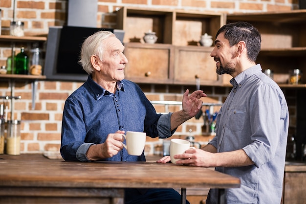 Pleasant elderly man talking to his father while drinking coffee in the kitchen