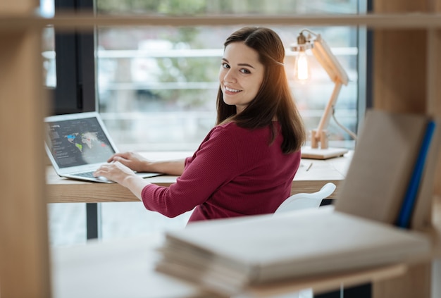 Pleasant emotions. Young lighthearted woman looking at you with a smile while sitting at the table and using a laptop