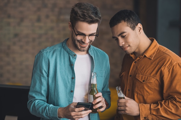 Pleasant happy man holding his smartphone while drinking beer with his friend