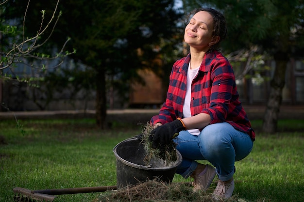 Pleasant housewife gardener enjoys gardening on sunny day puts mowed grass into bucket while cleaning the backyard for further composting