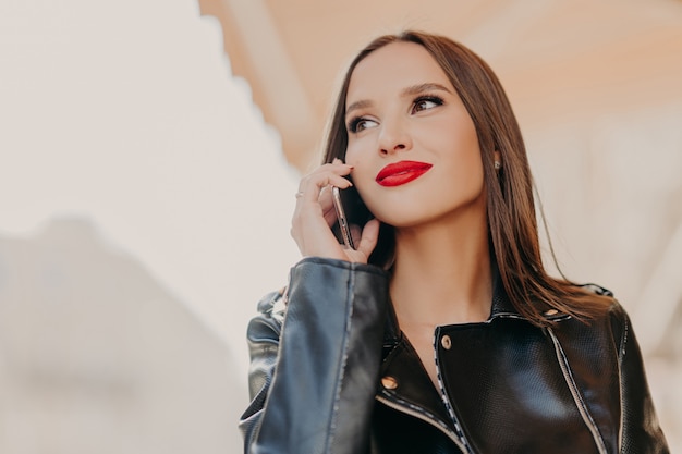 Pleasant looking dreamy woman focused aside, has telephone conversation, wears leather coat