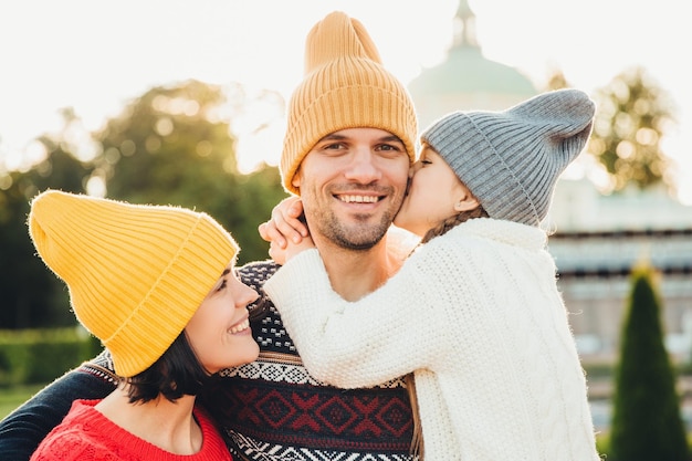 Pleasant looking little girl being thankful to her father for buying new bicycle kisses him in cheek Young family walk together outdoor spend spare time in park wear knitted fashionable hats