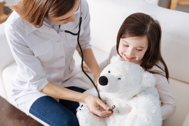 Pleasant loving mother examining with stethoscope fluffy toy of her daughter while pretending to be a doctor