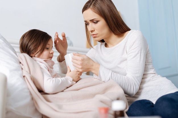 Pleasant loving mother taking care of her little sick daughter while sitting together on the sofa