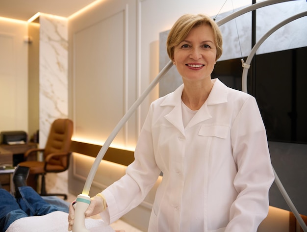 Pleasant mature female beautician smiling confidently looking at camera while doing body care procedures in spa clinic specializing in hardware cosmetology