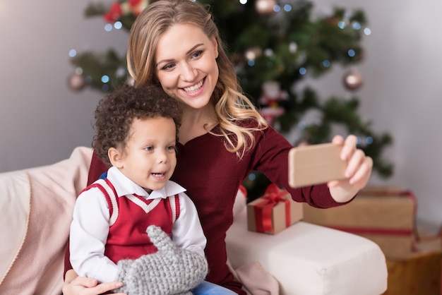 Pleasant memory. Beautiful delighted young woman sitting with her son of her smartphone while taking selfies