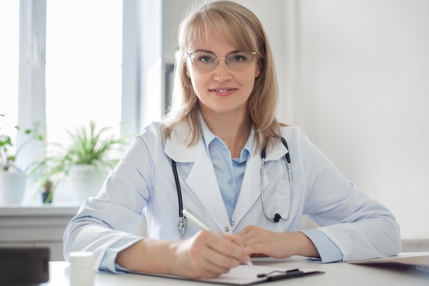 A pleasant middleaged blonde woman doctor in a white coat glasses with a stethoscope sits