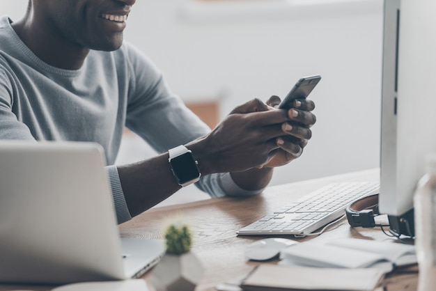 Pleasant moments. Close-up of handsome young African man typing a message on the phone and smiling while sitting at the desk in creative office