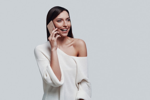 Pleasant talk. Attractive young woman talking on the smart phone and smiling while standing against grey background