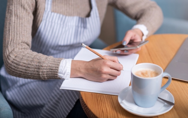 Pleasant work. Young pretty woman making notes and using cellphone while sitting by the table.