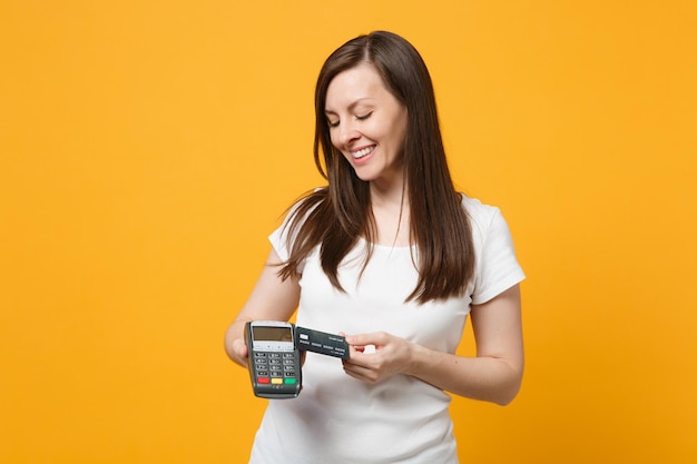 Pleasant young woman holding wireless modern bank payment terminal to process and acquire credit card payments isolated on yellow orange wall background. People lifestyle concept. Mock up copy space.