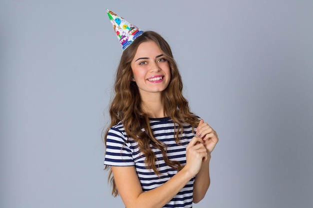 Pleasant young woman wearing in stripped T-shirt and celebration cap on grey background in studio