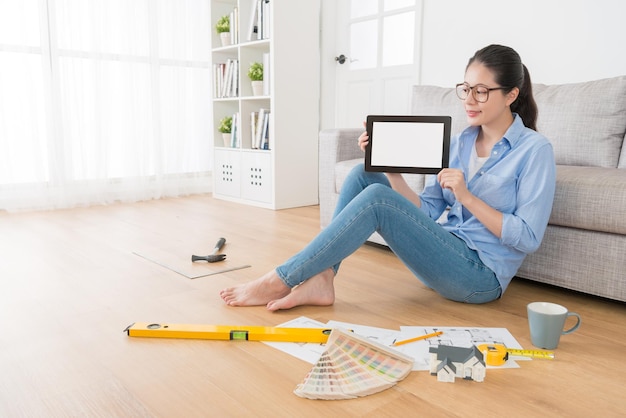 pleasantly attractive female college design department student doing homework in living room wooden floor and holding mobile pad computer showing blank screen.