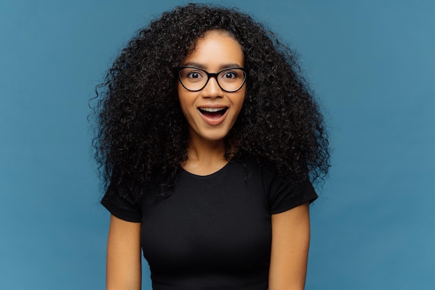 Pleasantly surprised dark skinned woman keeps jaw dropped gazes with interest has curly hair wears black t shirt models against blue wall Afro American lady gazes with shock has bated breath