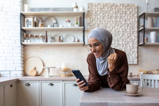 Pleasantly surprised young muslim woman in hijab at home in the kitchen looking at the phone with