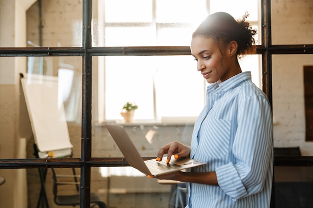 pleased african american woman smiling and typing on laptop while standing in modern office