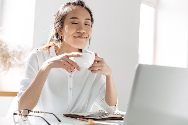 Pleased asian business woman drinking coffee and enjoys with closed eyes while sitting by the table in office