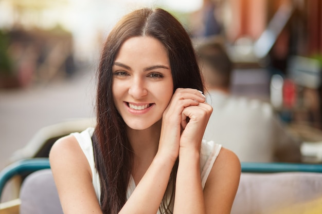 Pleased attractive lady, smiles gently at camera, enjoys spare time in outdoor cafeteria, rests at cozy place, being in good mood. Lovely woman has dinner break at sidewalk restaurant after work