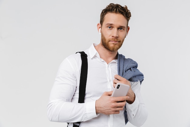 pleased blonde man using cellphone and wireless earphones isolated over white wall