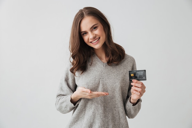 Pleased brunette woman in sweater holding credit card and pointing on it  over grey wall