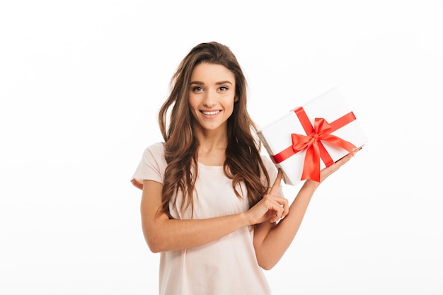 Pleased brunette woman in t-shirt holding gift box  over white wall